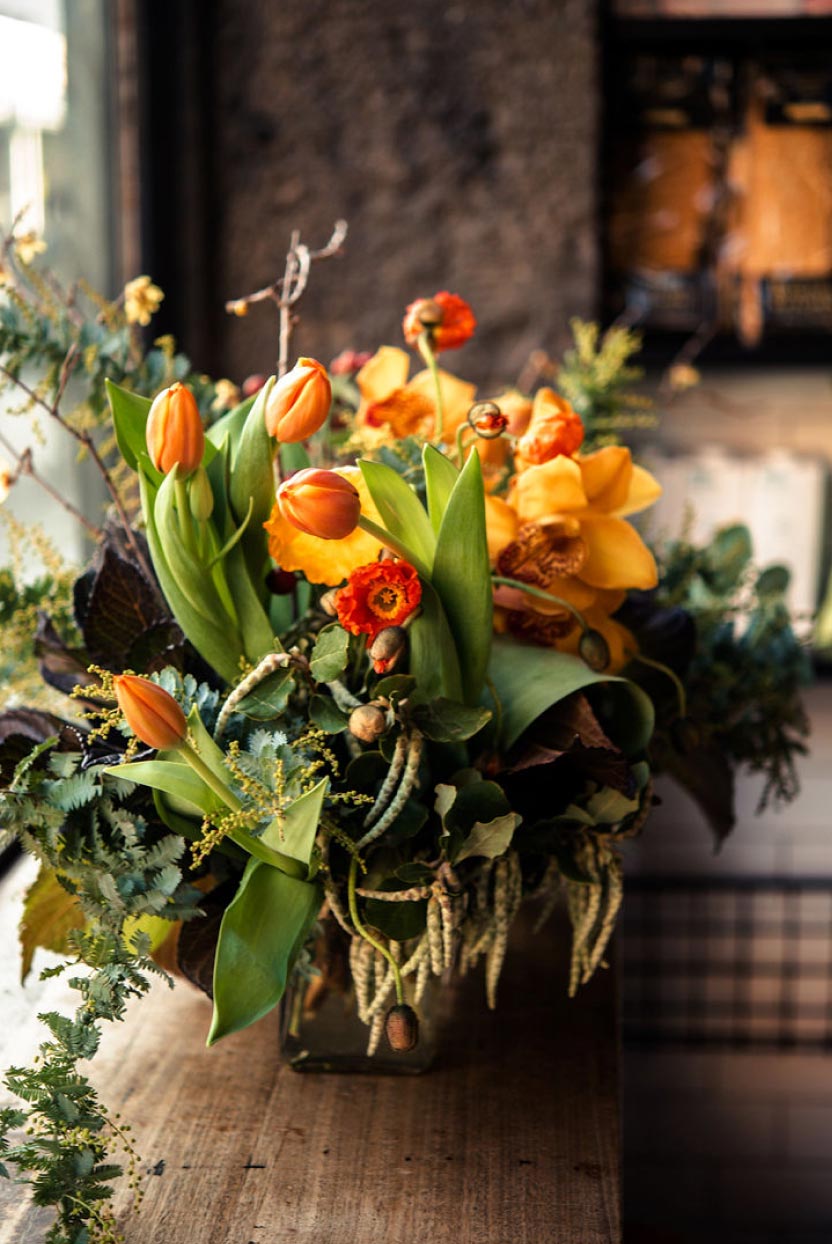 This photograph depicts a bouquet of flowers in a vase. The vase contains orange and red flowers and is sitting on a wooden table.