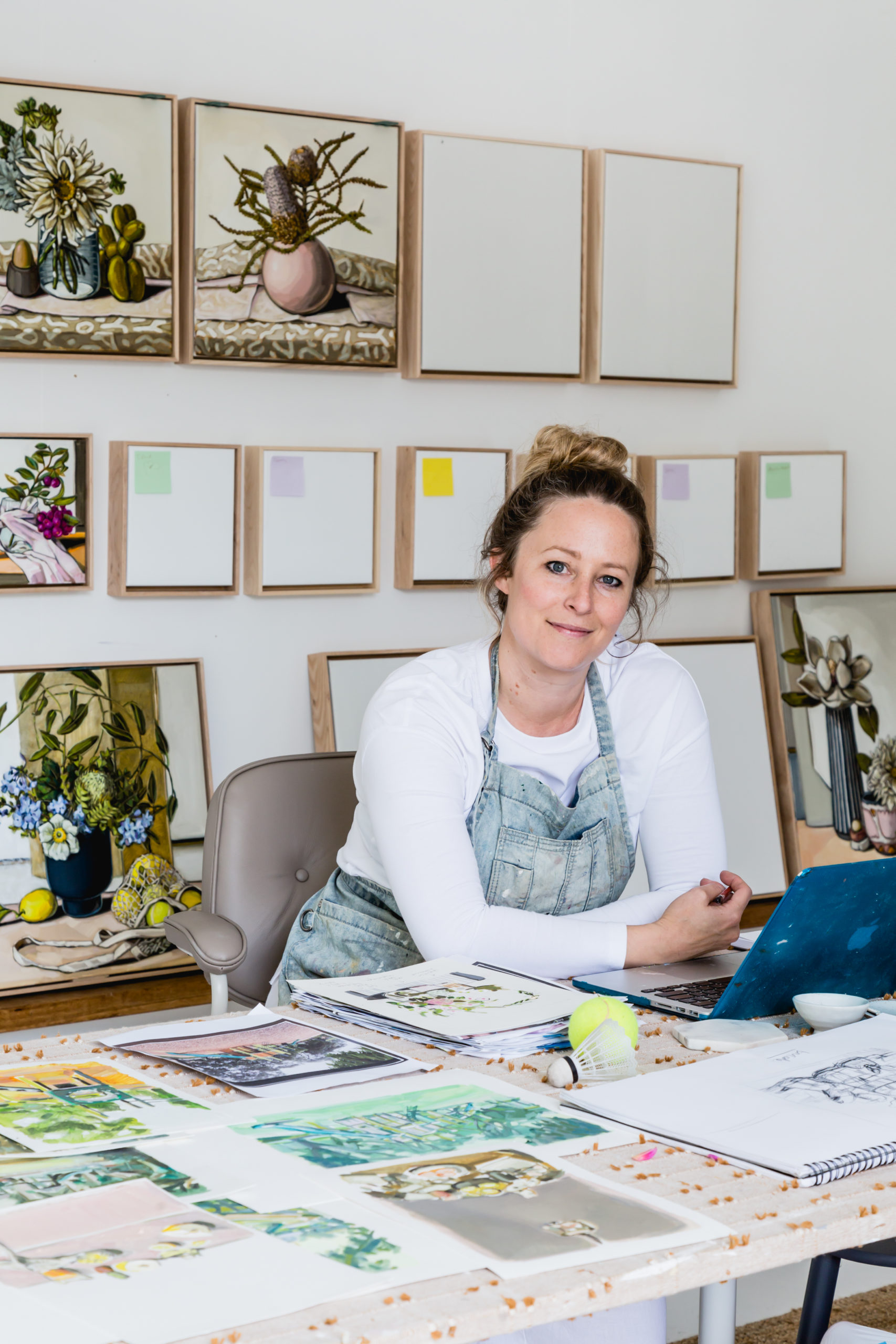 A photo of artist Sam Michelle on her laptop, surrounding by paintings of flowers.