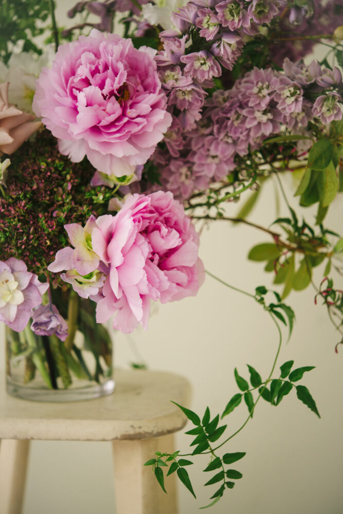 This photograph depicts a vase of flowers sitting on a stool. The flowers are pink in colour and contain peonies and delphinium.