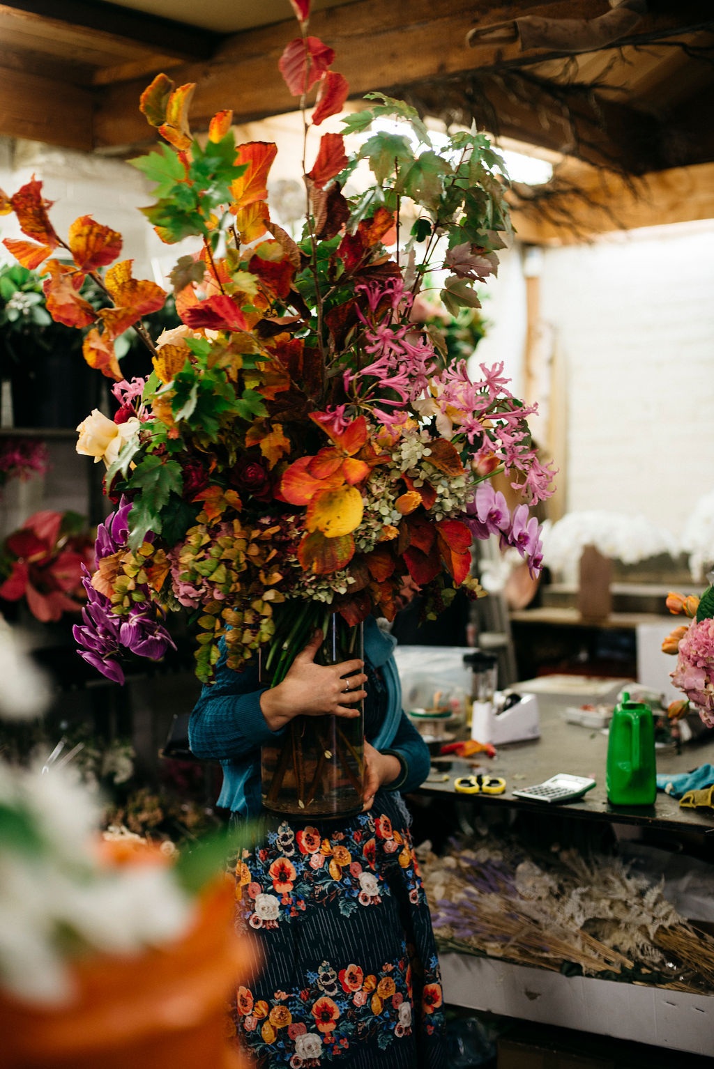 This photograph depicts a woman holding a very large glass vase of flowers. The flowers are brightly coloured pink, orange and purple. The vase also contains branches of orange and green leaves.