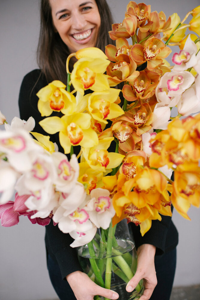 This photograph depicts a woman smiling and holding a vase of cymbidium orchids. The flowers are white, yellow and orange.