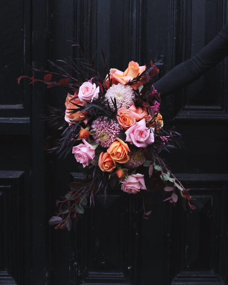 This photograph depicts a hand holding a bouquet of pink and peach coloured flowers. The background is a door painted black. The bouquet contains dark red coloured leaves.