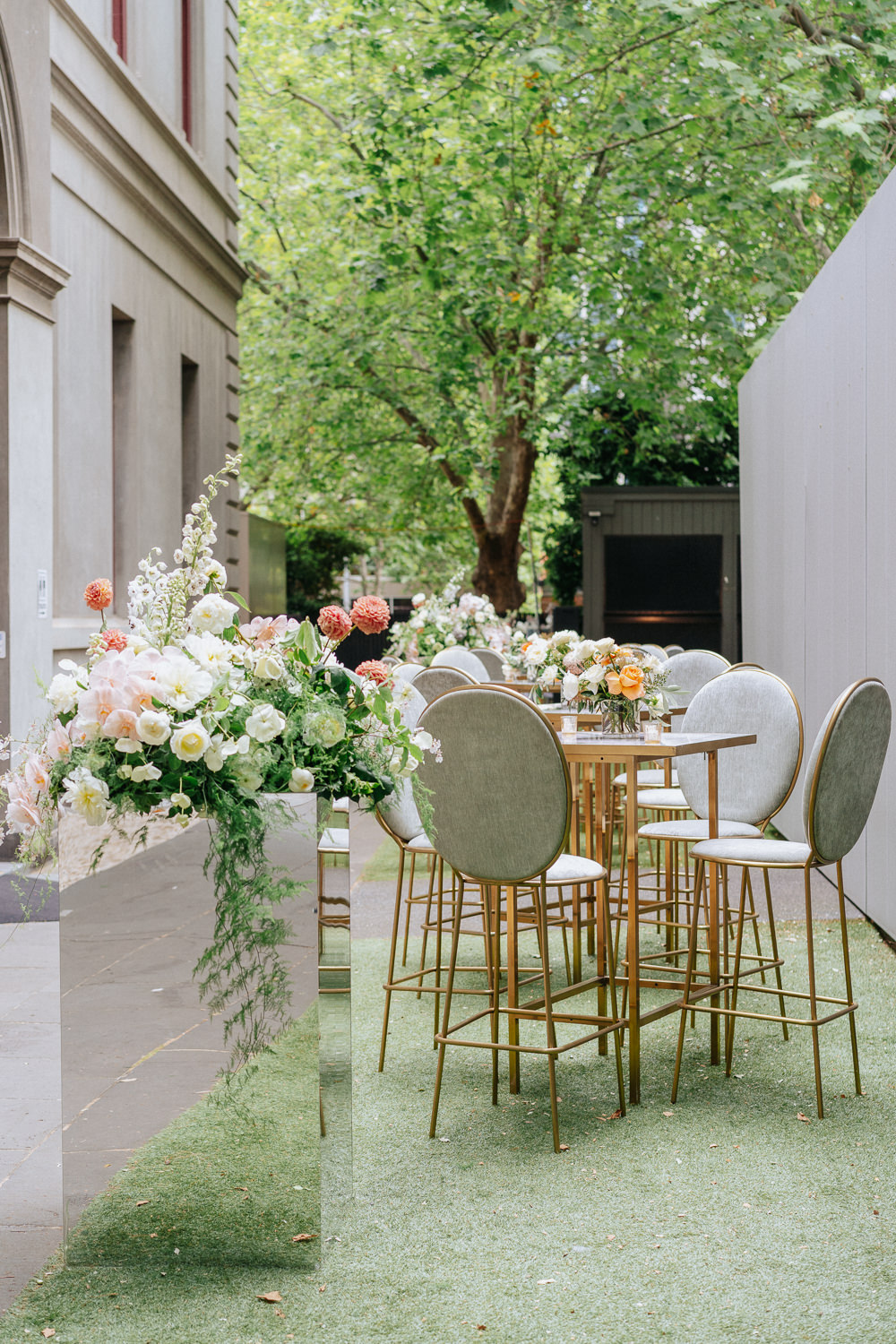 A pink, white and green floral arrangement on top of a mirrored stand. Beside the stand are some tall bar seats and tables. Each table has a small flower arrangement on it.