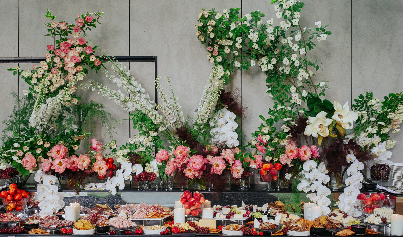 A buffet of food with a collection of floral arrangements behind it. Some of the flowers reach more than 1 metre above the table.