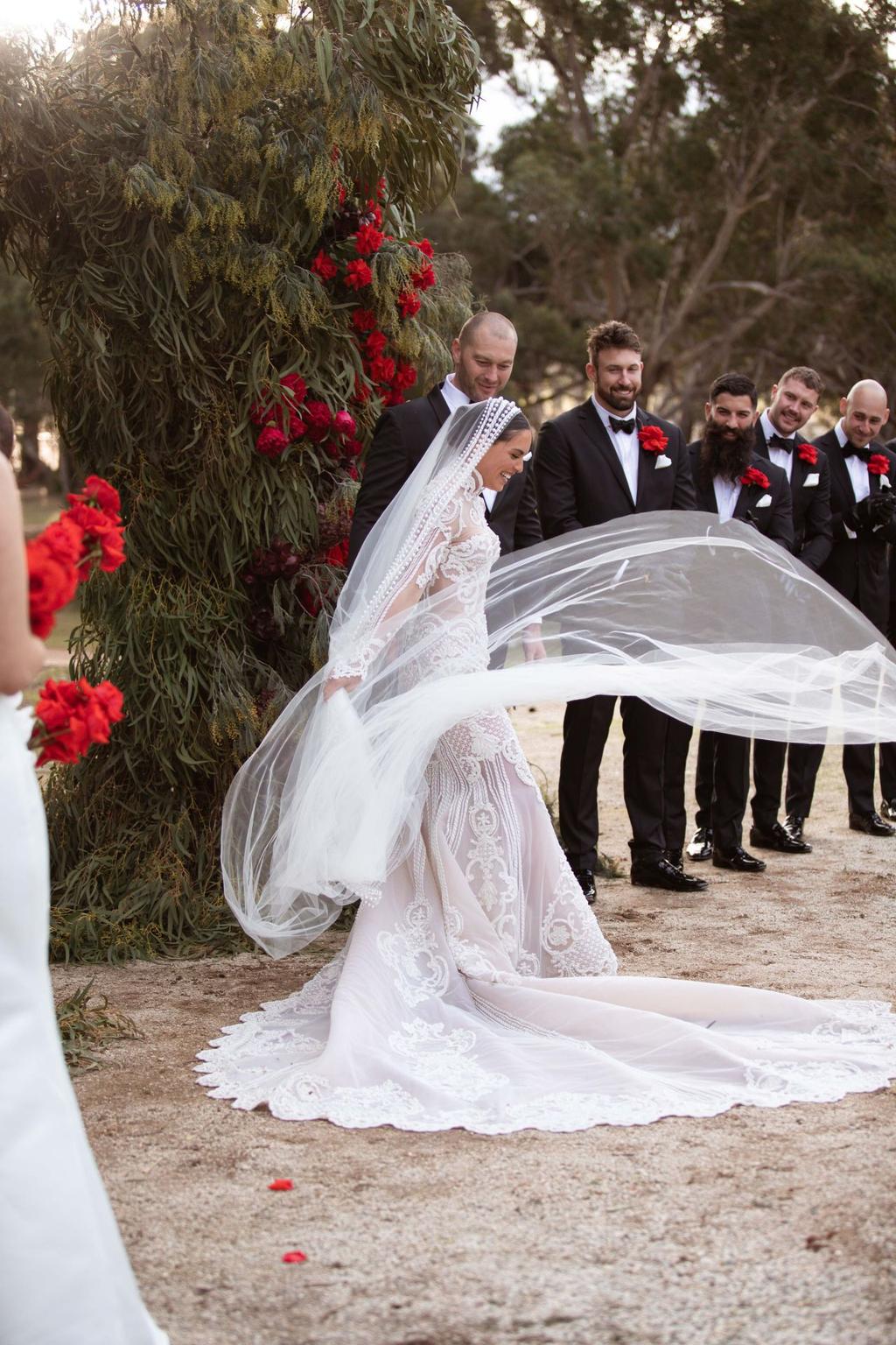 This photo depicts Olympia Valance at her wedding. A green and red floral arbor is behind her.