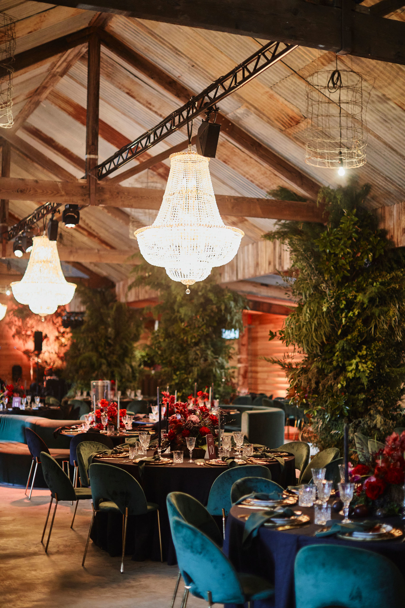 This photo depicts the inside of a barn, with crystal chandeliers and green velvet seats at dining tables. In the centre of each dining table is a deep red floral arrangement.