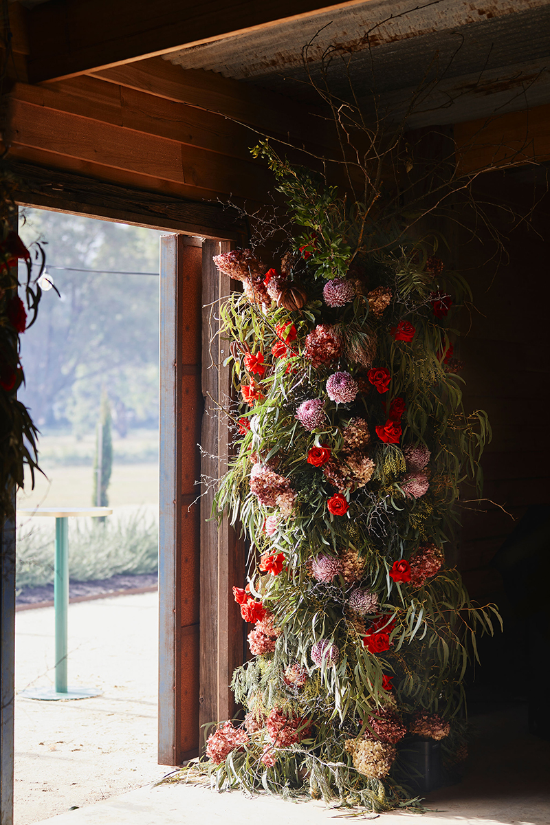A floral installation on the doorway of a barn. The foliage is native Australian leaves, the flowers are pale pink and bright red.