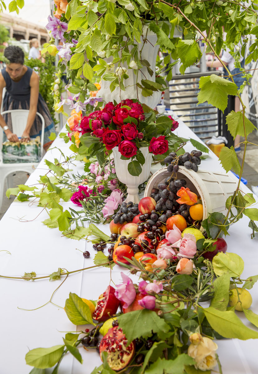 A table full of food and flowers. A vase of deep red roses in a white urn sits in the middle.