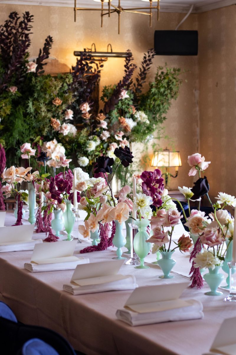 A dining table, with a collection of small green vases lined up in the centre. The vases contain red, pale pink, white and black flowers.