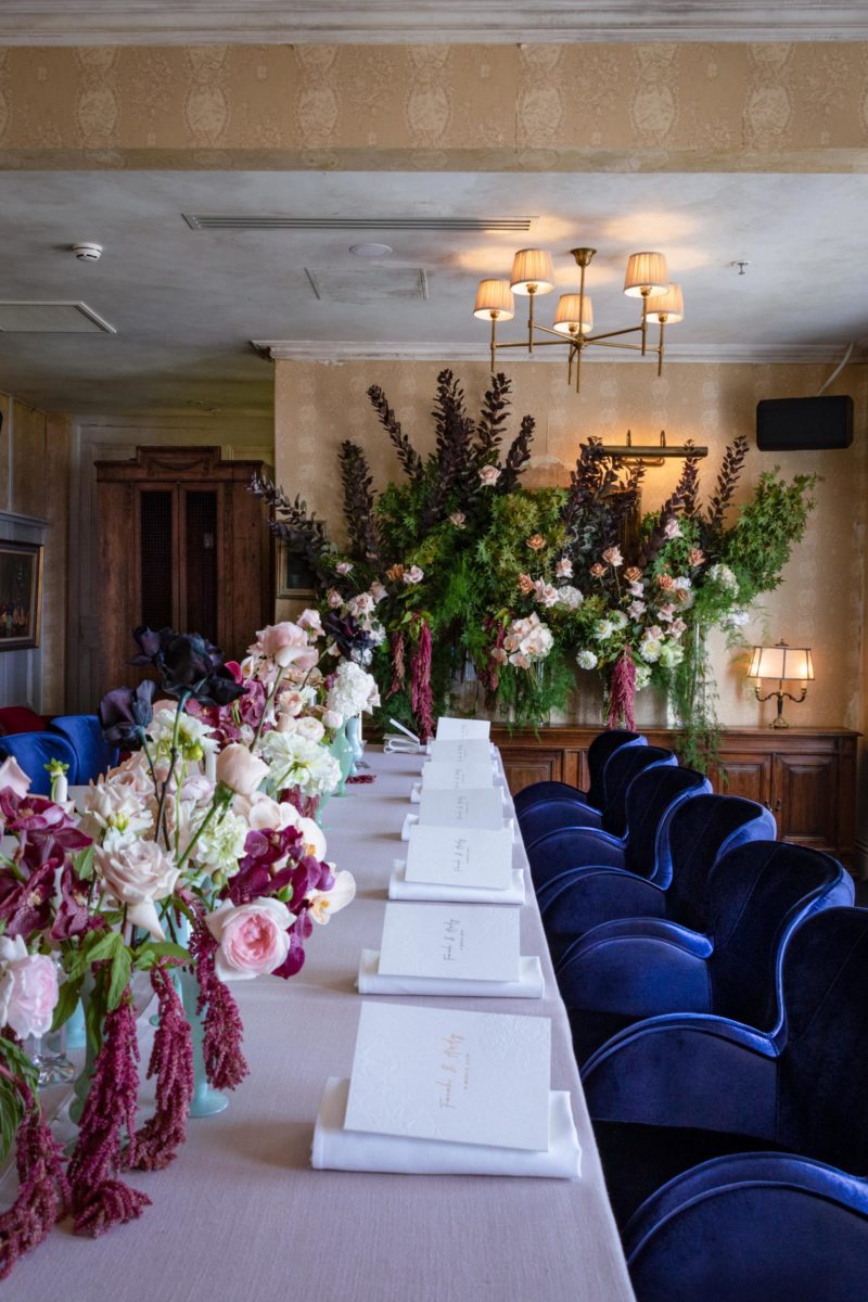 A dining table, with a collection of small green vases lined up in the centre. The vases contain red, pale pink, white and black flowers. Behind the table, several tall glass vases are lined up on a sideboard.