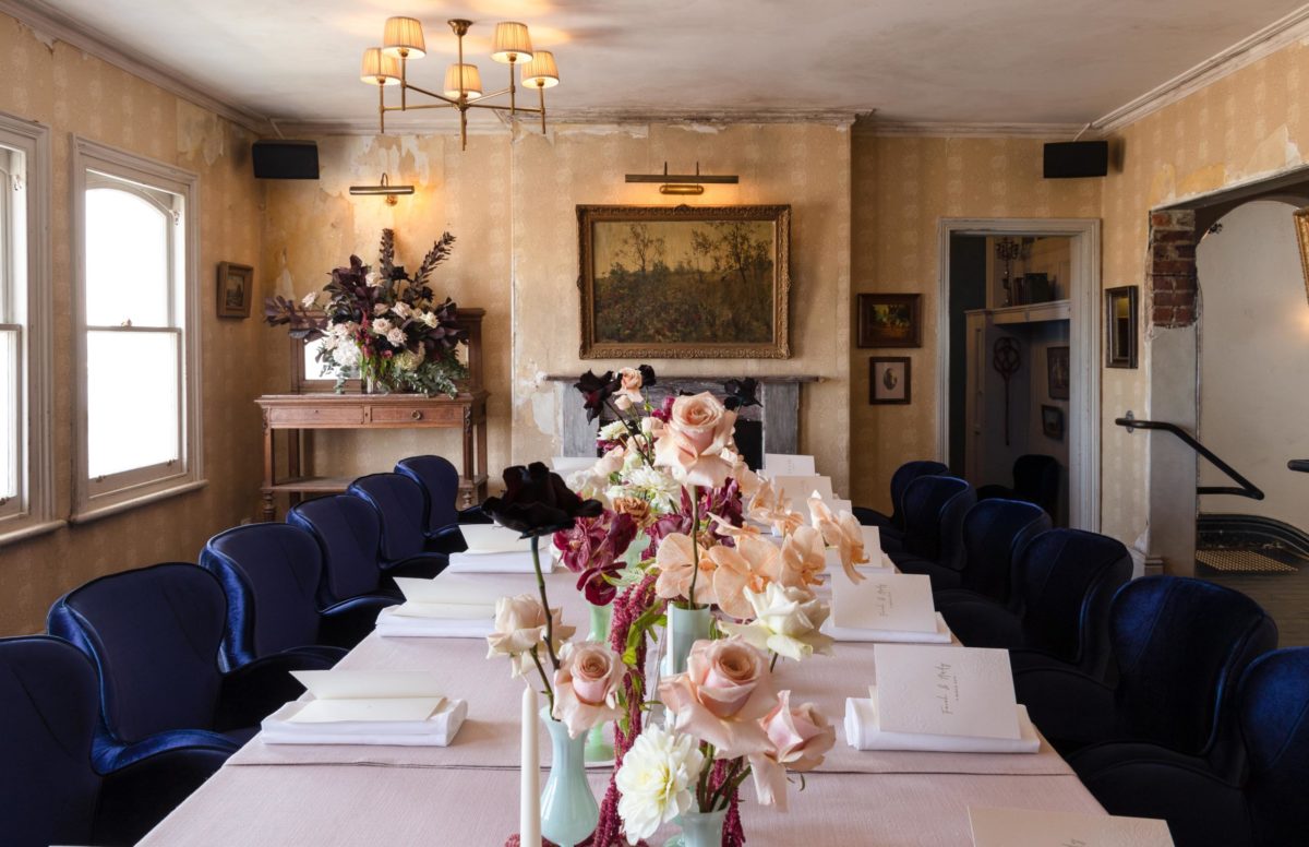 A dining table, with a collection of small green vases lined up in the centre. The vases contain red, pale pink, white and black flowers.