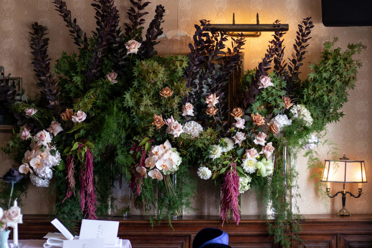 Several tall glass vases holding floral arrangements lined up against a wall. The flowers are pink, white and deep red. The foliage is green and dark purple.
