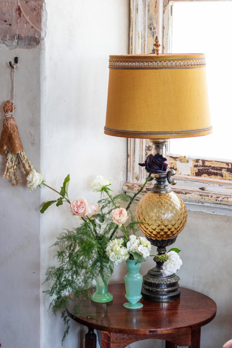 A lamp and two green vases on a small end table. The flowers are pink, white and green.