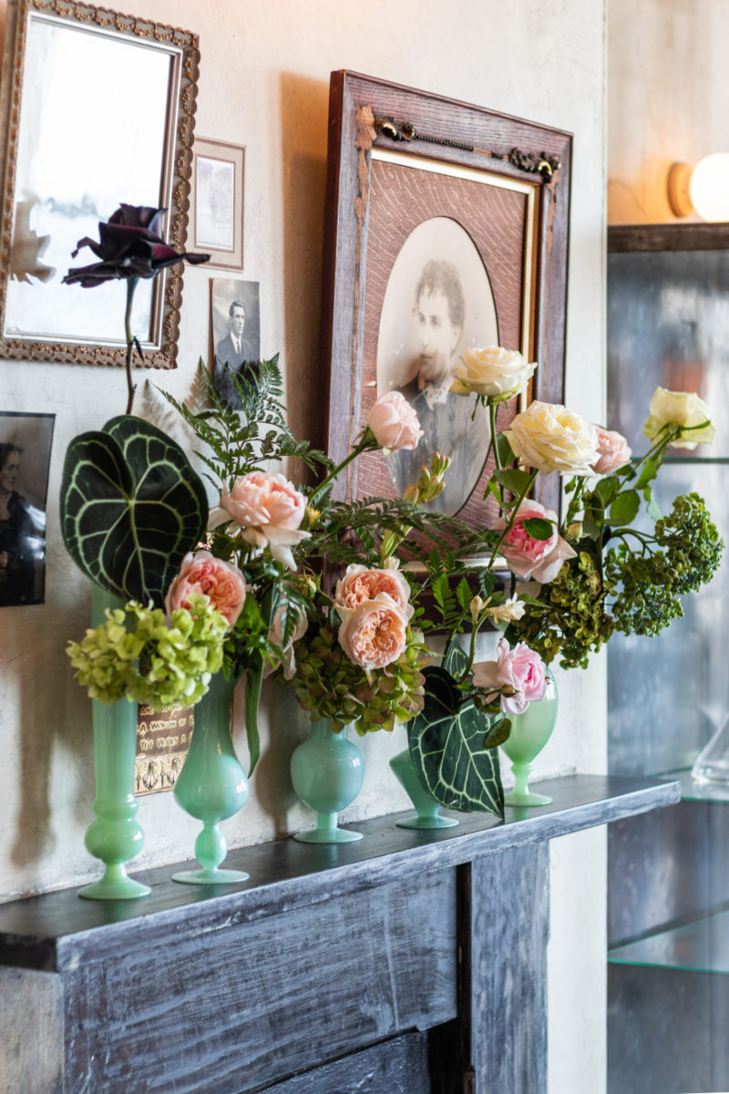 A set of jade green uniquely shaped vases, holding pink, white and green flowers. The vases are sitting on a mantle in a house.